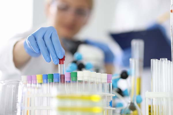 photo of a rack of test tubes with different colored caps, with a gloved hand inserting a tube into the rack; in the background, out of focus, the lab tech's face is slightly visible